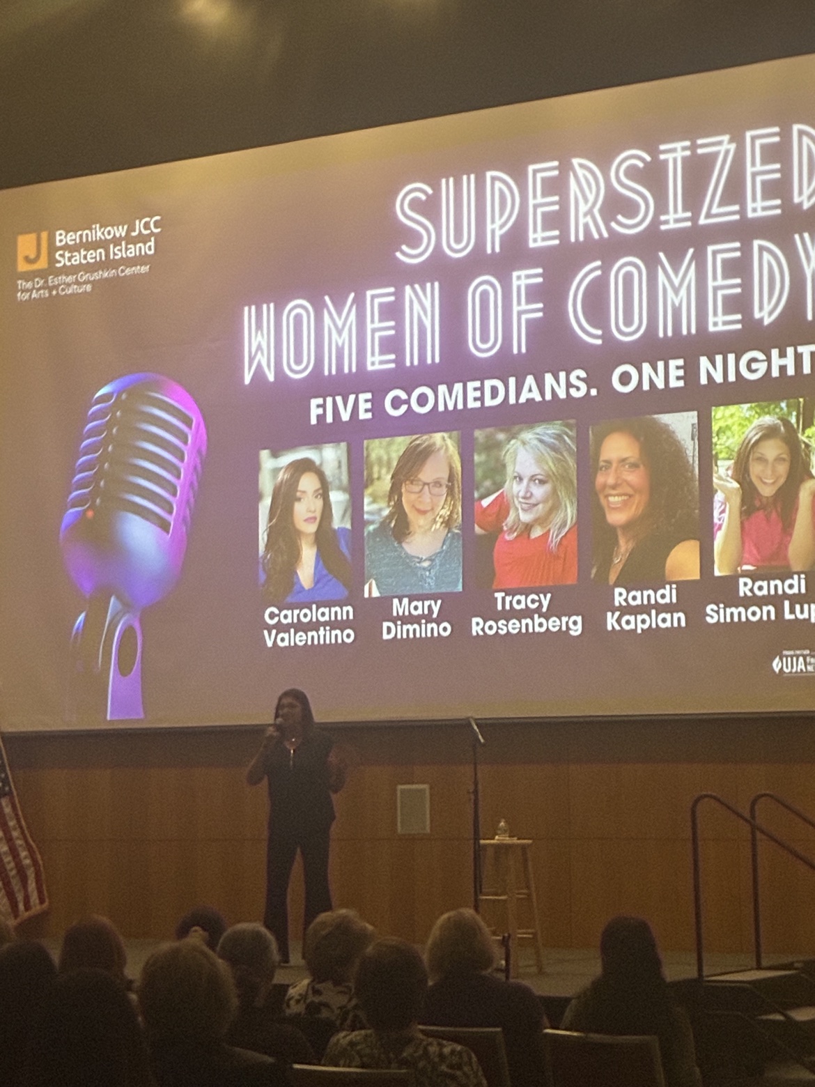 A comedian performing at the Grushkin Center with the Supersized Women of Comedy projection behind her
